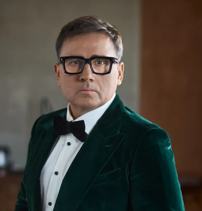 Andrzej Giza w zielonej aksamitnej marynarce, białej koszuli i czarnej muszce, stoi we wnętrzu sali koncertowej i patrzy w obiektyw.